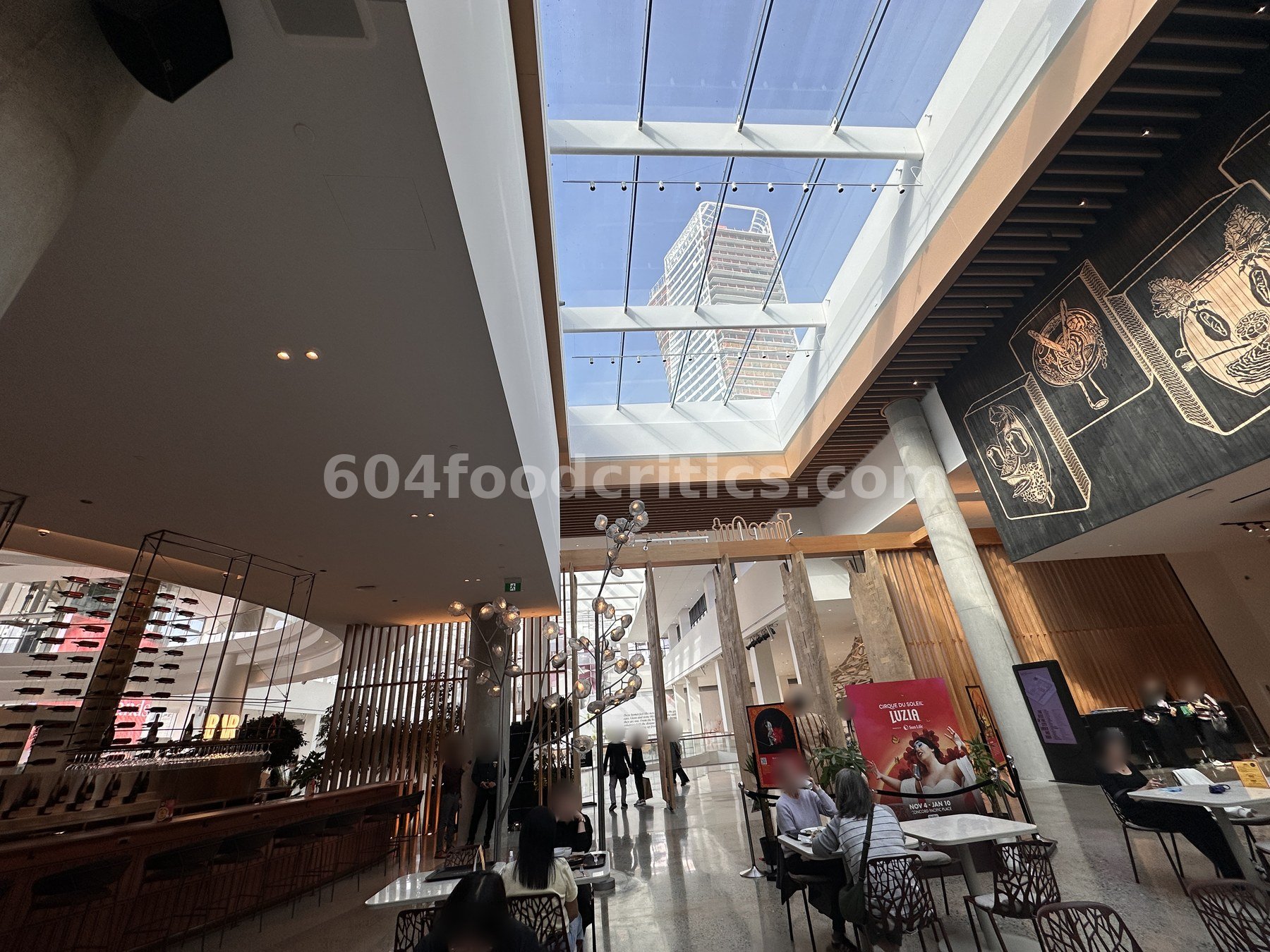 Dining hall at Time Out Market Vancouver in Oakridge Park under a large skylight, with diners, a bar and wood columns